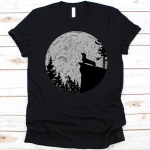 May include: Black t-shirt featuring a silhouette of a dachshund dog on a cliff, howling at a full moon. The moon is detailed with craters and surrounded by silhouetted trees. A simple, yet striking design.