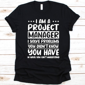 以下が含まれることがあります： 「I AM A PROJECT MANAGER I SOLVE PROBLEMS YOU DIDN'T KNOW YOU HAVE IN WAYS YOU CAN'T UNDERSTAND」と白い文字で書かれた黒いTシャツ。Tシャツはクルーネックで半袖です。