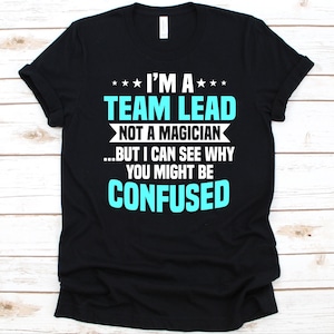 Puede incluir: Camiseta negra con el texto "I'M A TEAM LEAD NOT A MAGICIAN...BUT I CAN SEE WHY YOU MIGHT BE CONFUSED" en blanco y turquesa. El texto es en negrita y claro.