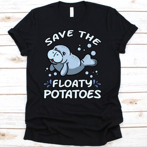 Puede incluir: Camiseta negra con un dibujo de manatí de dibujos animados y el texto "SAVE THE FLOATY POTATOES". El manatí es de color azul claro y sonríe, con burbujas y salpicaduras de agua a su alrededor. La camiseta es de material suave.