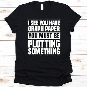 Puede incluir: Camiseta negra con texto blanco: "VEO QUE TIENES PAPEL CUADRICULADO, DEBES ESTAR TRAMANDO ALGO". La camiseta tiene un diseño de papel cuadriculado verde detrás del texto.