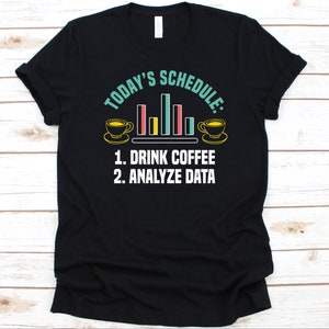 May include: Black t-shirt featuring the text "TODAY'S SCHEDULE: 1. DRINK COFFEE 2. ANALYZE DATA." The design includes coffee cups and a bar graph in yellow, red, and blue. The text and graphics are in white and teal.