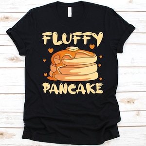 May include: Black t-shirt featuring a stack of pancakes with syrup and butter, and the words "Fluffy Pancake" in a playful font. Small orange hearts are scattered around the design. A casual and comfortable tee.