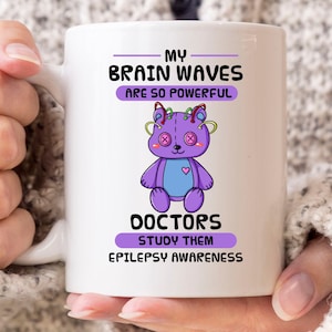 May include: White ceramic mug with a purple teddy bear design and the text "MY BRAIN WAVES ARE SO POWERFUL" and "DOCTORS STUDY THEM EPILEPSY AWARENESS". The mug has a curved handle.