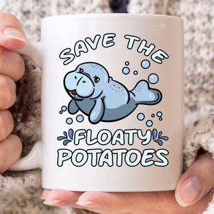 Puede incluir: Taza de cerámica blanca con una ilustración de manatí de dibujos animados y el texto "SAVE THE FLOATY POTATOES". El manatí es azul claro, sonriente, con burbujas y salpicaduras de agua. La taza tiene un asa blanca.