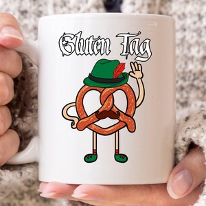 Op de afbeelding: Witte keramische mok met een cartoon pretzel karakter met een groene hoed en snor, die zwaait. De mok heeft de woorden "Gluten Tag". De pretzel draagt groene schoenen en rood-wit gestreepte sokken. Een leuk, nieuwigheid artikel.