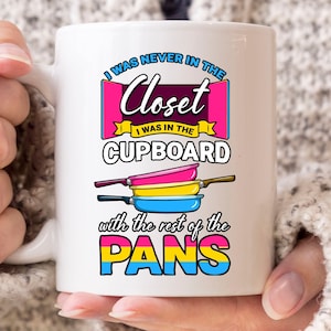 May include: White ceramic mug with the text "I WAS NEVER IN THE Closet I WAS IN THE CUPBOARD with the rest of the PANS" in a colorful design. The design includes a pink, yellow, and blue color scheme with a pansexual pride flag.