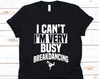Puede incluir: Camiseta negra con texto blanco que dice "I CAN'T I'M VERY BUSY BREAKDANCING." Una silueta blanca de un bailarín de breakdance está debajo del texto. La camiseta está hecha de un material suave.