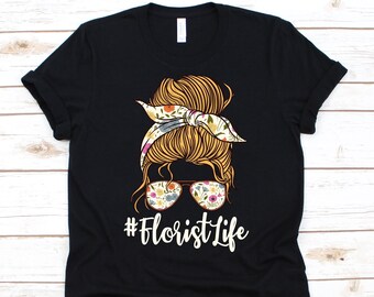 May include: Black t-shirt with a floral design. The graphic shows a woman's hairstyle with a floral headband and sunglasses. The text "#Florist Life" is printed in white below the graphic. The shirt is made of a soft material.