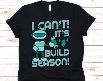 May include: Black t-shirt with a teal and white robot graphic holding a wrench. The text reads "I CAN'T! IT'S BUILD SEASON!" in teal and white. The shirt is made of a soft material.