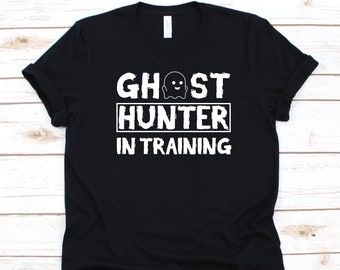 Może przedstawiać: Czarny t-shirt z białym napisem "GHOST HUNTER IN TRAINING". Tekst jest ułożony w trzech linijkach, z grafiką ducha nad słowem "HUNTER". Koszulka ma krótkie rękawy i jest wykonana z miękkiego materiału.