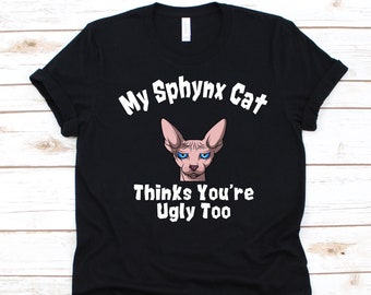 Może przedstawiać: Czarna koszulka z białym napisem "My Sphinx Cat Thinks You're Ugly Too". Grafika przedstawia kreskówkową ilustrację kota Sfinksa z niebieskimi oczami i ponurym wyrazem twarzy.