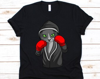 Puede incluir: Camiseta negra con un gato boxeador de dibujos animados. El gato es gris con ojos verdes y lleva una bata con capucha negra y guantes de boxeo rojos. El gato está en posición de boxeo.