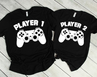 May include: Two black t-shirts with white graphics. One shirt says "PLAYER 1" above a white controller graphic. The other shirt says "PLAYER 2" above a matching controller graphic. The shirts are likely made of cotton or a similar fabric.