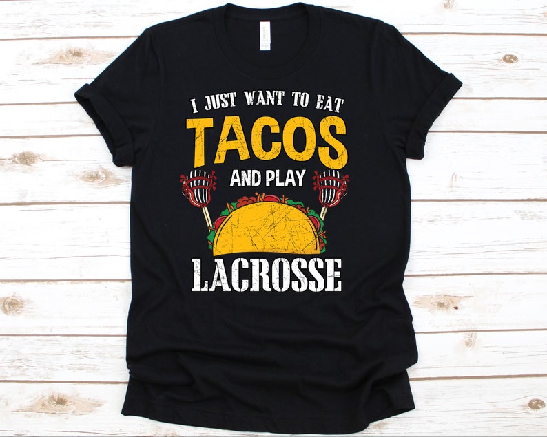 May include: Black t-shirt with the text "I JUST WANT TO EAT TACOS AND PLAY LACROSSE" in yellow and white. A graphic of a taco and lacrosse sticks is also featured on the shirt.
