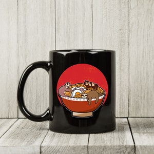 May include: Black ceramic mug featuring a cartoon illustration of a bowl of ramen with sloths. The ramen bowl is red with a white rim and contains noodles, an egg, and several sloths. A red circle is behind the bowl.