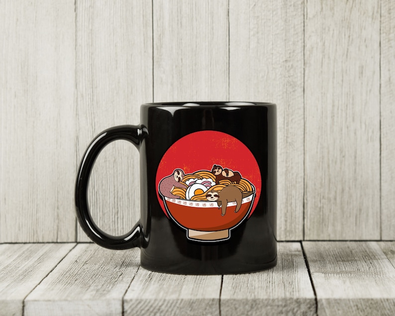 May include: Black ceramic mug featuring a cartoon illustration of a bowl of ramen with sloths. The ramen bowl is red with a white rim and contains noodles, an egg, and several sloths. A red circle is behind the bowl.