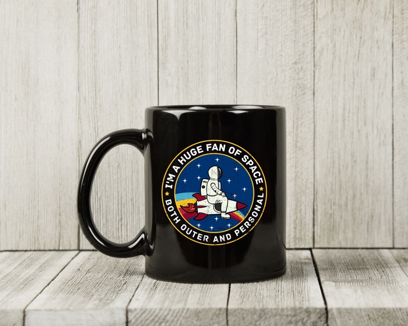 May include: Black ceramic mug with a circular graphic featuring an astronaut riding a rocket. The graphic includes the text "I'M A HUGE FAN OF SPACE BOTH OUTER AND PERSONAL" in a blue and yellow design. The mug has a large handle.