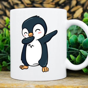 May include: White ceramic mug featuring a cartoon penguin in a dabbing pose. The penguin is black and white with an orange beak and pink cheeks. The mug has a curved handle and is set against a blurred background of greenery.