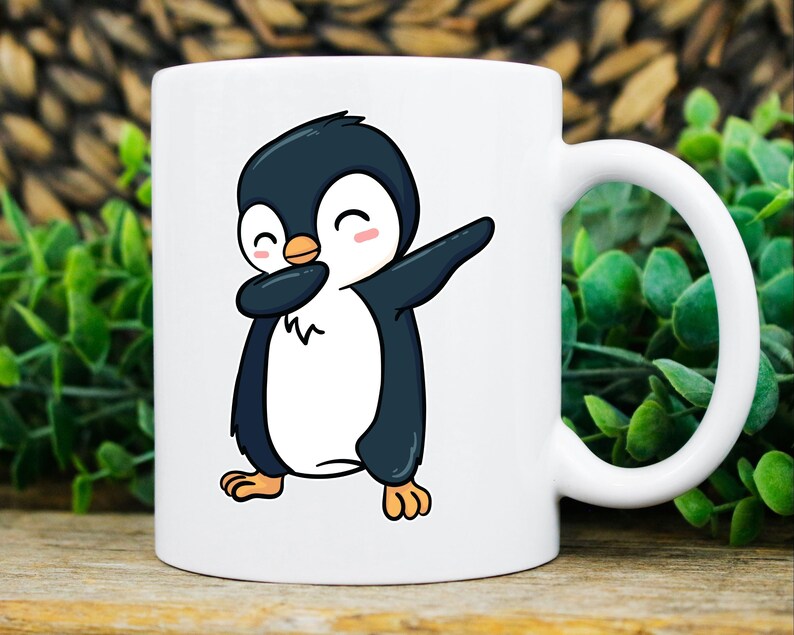 May include: White ceramic mug featuring a cartoon penguin in a dabbing pose. The penguin is black and white with an orange beak and pink cheeks. The mug has a curved handle and is set against a blurred background of greenery.