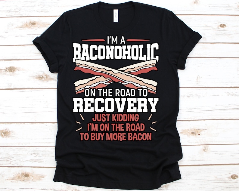 May include: Black t-shirt with the text "I'M A BACONOHOLIC ON THE ROAD TO RECOVERY JUST KIDDING I'M ON THE ROAD TO BUY MORE BACON". The design includes illustrations of bacon strips in shades of brown and white.