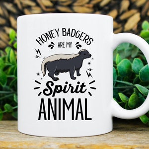 May include: White ceramic mug with a black and gray honey badger illustration. The text on the mug reads "HONEY BADGERS ARE MY Spirit ANIMAL" in a black, bold font. The mug has a white handle and is set against a blurred background.