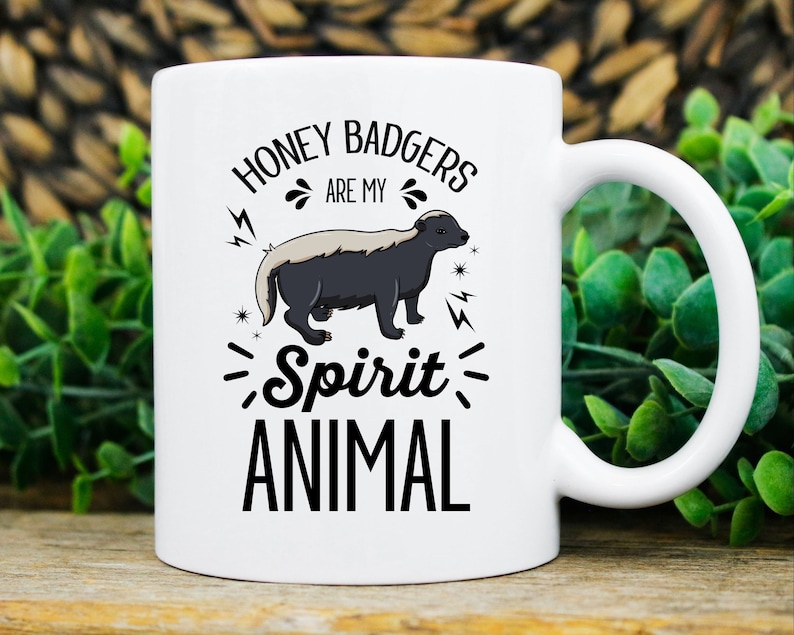 May include: White ceramic mug with a black and gray honey badger illustration. The text on the mug reads "HONEY BADGERS ARE MY Spirit ANIMAL" in a black, bold font. The mug has a white handle and is set against a blurred background.
