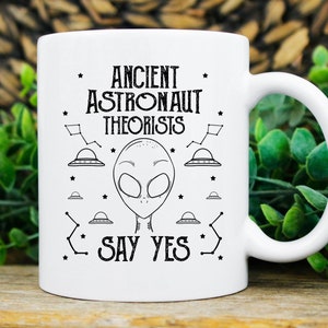 Puede incluir: Taza de cer&aacute;mica blanca con un dise&ntilde;o gr&aacute;fico negro que presenta una cabeza alien&iacute;gena, ovnis y el texto "ANCIENT ASTRONAUT THEORISTS SAY YES". La taza tiene un asa curva y es adecuada para bebidas fr&iacute;as o calientes.