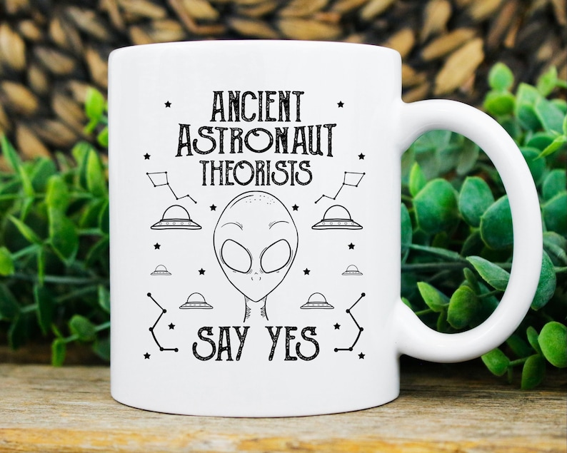 Puede incluir: Taza de cer&aacute;mica blanca con un dise&ntilde;o gr&aacute;fico negro que presenta una cabeza alien&iacute;gena, ovnis y el texto "ANCIENT ASTRONAUT THEORISTS SAY YES". La taza tiene un asa curva y es adecuada para bebidas fr&iacute;as o calientes.