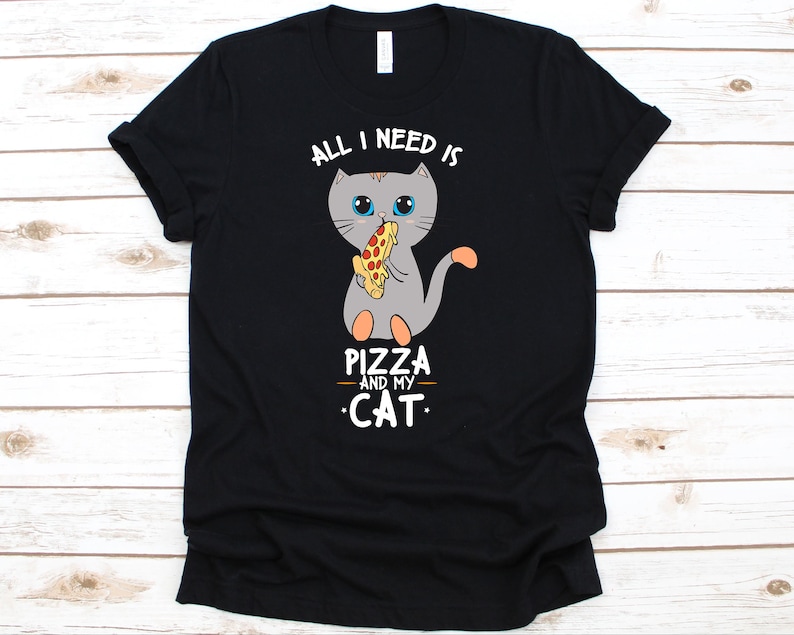 May include: Black t-shirt featuring a gray cartoon cat eating pizza. The text "ALL I NEED IS PIZZA AND MY CAT" is above and below the cat illustration. The shirt is made of a soft material.