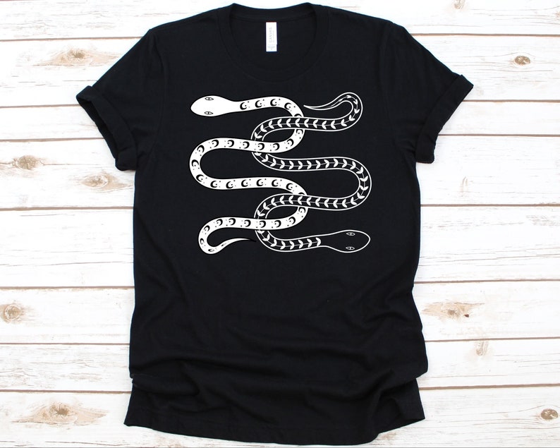 K&ouml;nnte beinhalten: Schwarzes T-Shirt mit einem wei&szlig;en Schlangenmuster. Die Schlange ist mit Halbmond- und Pfeilmuster verziert. Das Design ist mittig auf dem T-Shirt platziert. Das T-Shirt hat einen Rundhalsausschnitt und kurze &Auml;rmel.