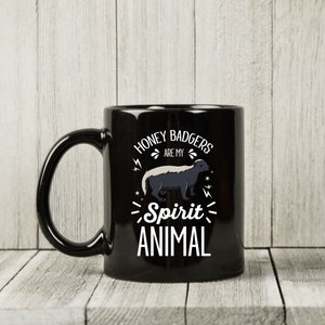 May include: Black ceramic mug with a white graphic of a honey badger and the text "Honey Badgers are my Spirit Animal". The mug has a large handle and is set against a light wood background.