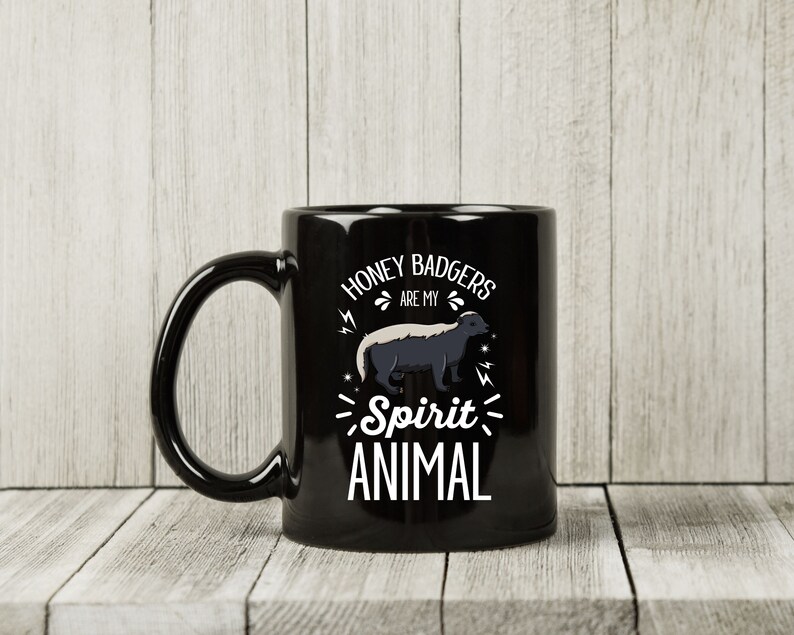 May include: Black ceramic mug with a white graphic of a honey badger and the text "Honey Badgers are my Spirit Animal". The mug has a large handle and is set against a light wood background.