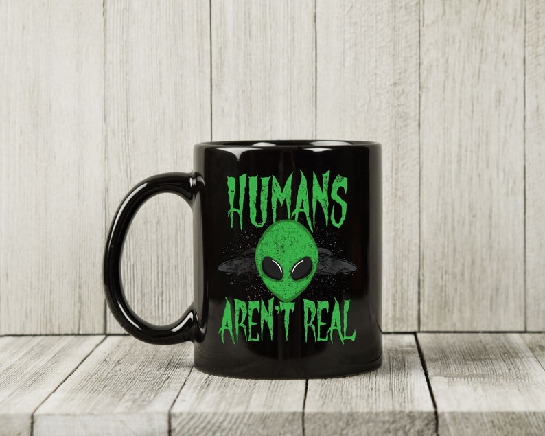 May include: Black ceramic mug with a green alien face graphic and the text "HUMANS AREN'T REAL". The alien has large black eyes and is set against a starry background. The mug has a large, curved handle.