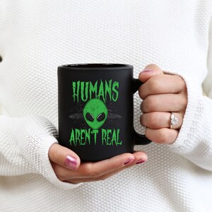 May include: Black ceramic mug with the text "HUMANS AREN'T REAL" in green, featuring a green alien head graphic. The mug has a black handle and a matte finish. The mug is held by a person wearing a white sweater.