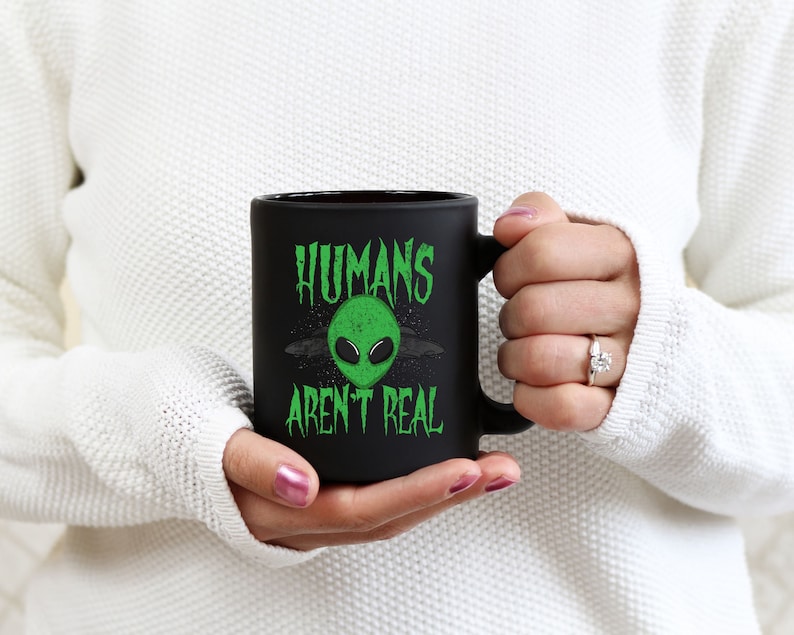 May include: Black ceramic mug with the text "HUMANS AREN'T REAL" in green, featuring a green alien head graphic. The mug has a black handle and a matte finish. The mug is held by a person wearing a white sweater.