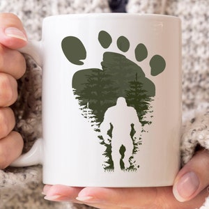 May include: White ceramic mug featuring a Bigfoot design. The design includes a large green footprint, a forest scene, and a silhouette of Bigfoot. The mug has a curved handle and is held by a person.