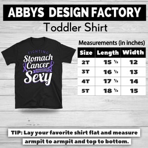 Op de afbeelding: Zwart peuter T-shirt met de tekst "FIGHTING Stomach Cancer AND STILL THIS Sexy" in wit en paars. Afmetingen in inches voor maten 2T tot 5T. Het shirt is van Abbys Design Factory.