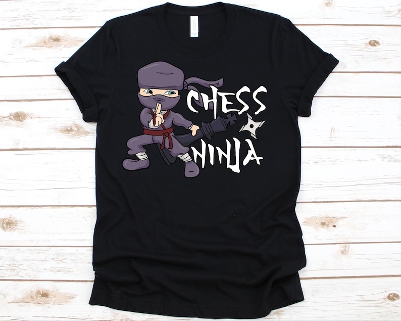 May include: Black t-shirt featuring a cartoon ninja with a chess piece and throwing star. The ninja is wearing a purple outfit and has the words "Chess Ninja" in white. The shirt is made of a soft material.