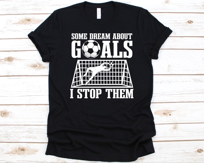 Puede incluir: Camiseta negra con un gr&aacute;fico blanco que representa un bal&oacute;n de f&uacute;tbol, una porter&iacute;a y un portero. El texto dice: "ALGUNOS SUE&Ntilde;AN CON GOLES, YO LOS PARO."