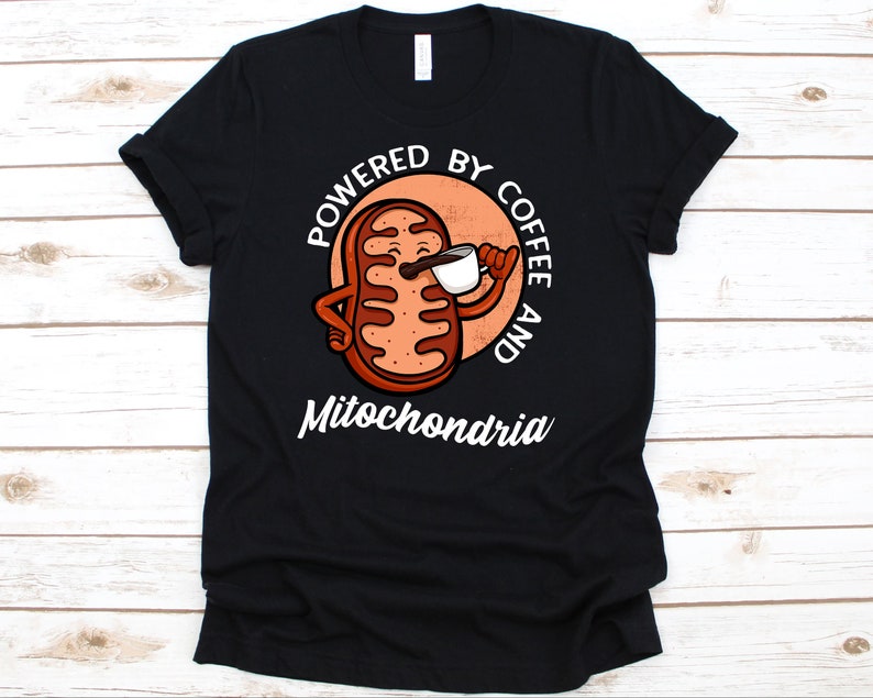 May include: Black t-shirt featuring a cartoon illustration of a mitochondria cell holding a coffee cup. The text reads "Powered by Coffee and Mitochondria" in white, with the word "Mitochondria" below the illustration. The design is set against a peach-colored circle.