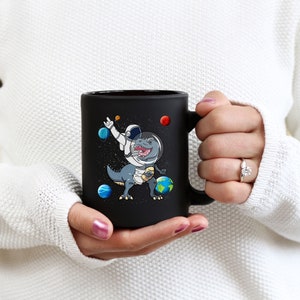 May include: Black ceramic mug featuring a cartoon dinosaur in an astronaut suit, surrounded by planets and stars. The dinosaur is dabbing with one arm raised. The mug is held by a person wearing a white sweater.