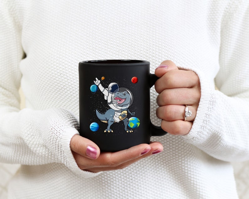 May include: Black ceramic mug featuring a cartoon dinosaur in an astronaut suit, surrounded by planets and stars. The dinosaur is dabbing with one arm raised. The mug is held by a person wearing a white sweater.