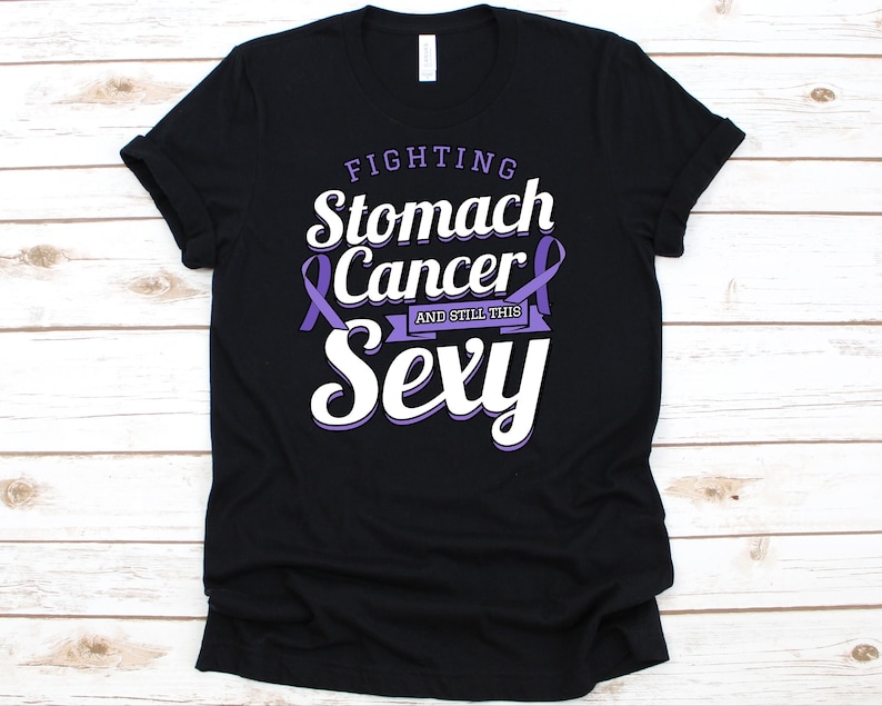 Op de afbeelding: Zwart T-shirt met de tekst "Fighting Stomach Cancer and Still This Sexy" in wit en paars. Het ontwerp bevat een paars bewustzijns lint. Het shirt is gemaakt van een zacht materiaal.