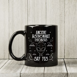 Puede incluir: Taza de cer&aacute;mica negra con el texto blanco "ANCIENT ASTRONAUT THEORISTS SAY YES". El dise&ntilde;o incluye una cara alien&iacute;gena, constelaciones y platillos volantes. La taza tiene un asa curva.