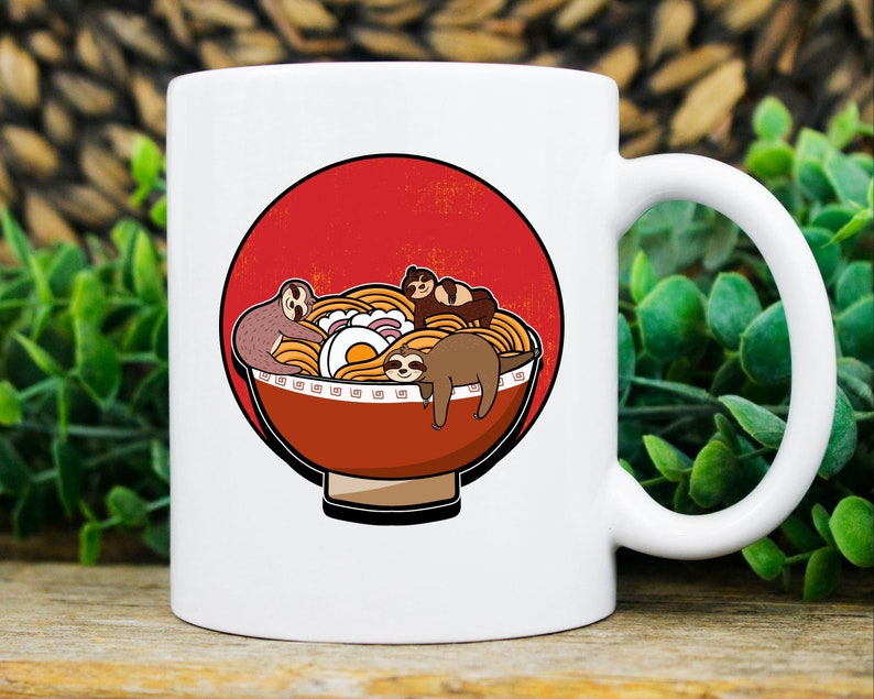 May include: White ceramic mug featuring a cartoon illustration of sloths lounging in a bowl of ramen noodles. The ramen bowl is red and brown, set against a red circle. The mug has a white handle and is perfect for coffee, tea, or any beverage.