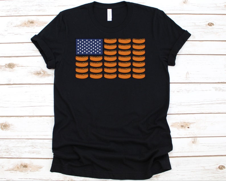 May include: Black t-shirt featuring a graphic of the American flag, with the stars replaced by white stars on a blue background and the stripes replaced by rows of hot dogs. The shirt has a crew neck and short sleeves.