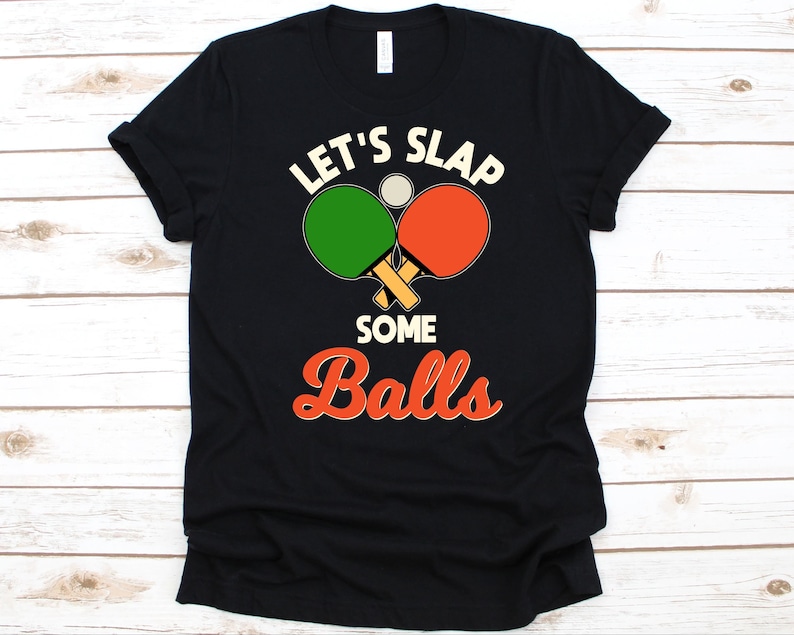 K&ouml;nnte beinhalten: Schwarzes T-Shirt mit einem grafischen Design, das zwei gekreuzte Tischtennisschl&auml;ger, einen gr&uuml;nen und einen orangefarbenen, sowie einen wei&szlig;en Tischtennisball zeigt. Der Text "LET'S SLAP SOME BALLS" ist oberhalb und unterhalb der Schl&auml;ger gedruckt.