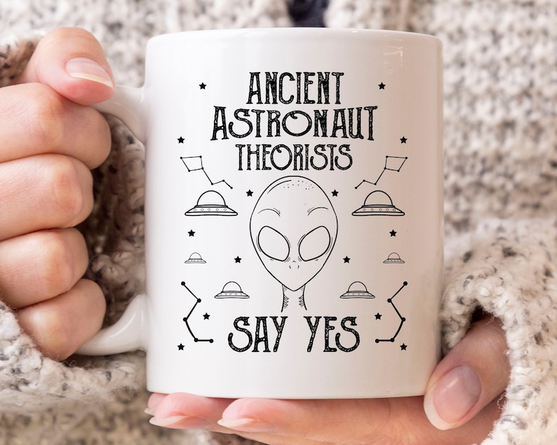 Puede incluir: Taza de cer&aacute;mica blanca con el texto negro "ANCIENT ASTRONAUT THEORISTS SAY YES", acompa&ntilde;ada de ilustraciones de una cabeza alien&iacute;gena, ovnis y constelaciones. La taza tiene un asa curvada.