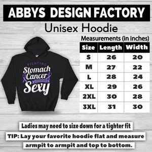 Op de afbeelding: Zwarte unisex hoodie met capuchon, met de tekst "FIGHTING Stomach Cancer AND STILL Sexy" in wit en paars. Maattabel met afmetingen in inches is inbegrepen. De merknaam "ABBYS DESIGN FACTORY" staat bovenaan.